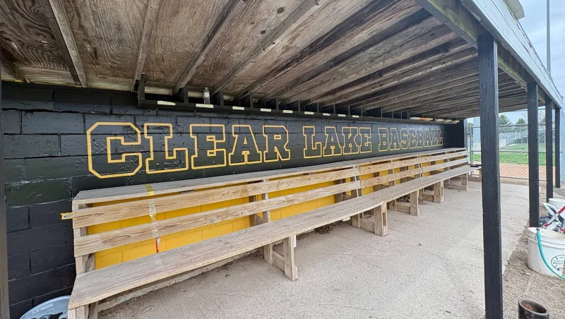 Baseball dugout graphics reading CLEAR LAKE BASEBALL in gold lettering on a black dugout wall above the team benches