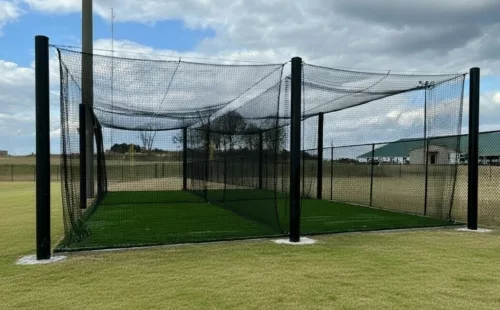 Outdoor baseball cage with black steel poles, cable-suspended netting, and artificial turf installed at a school baseball field