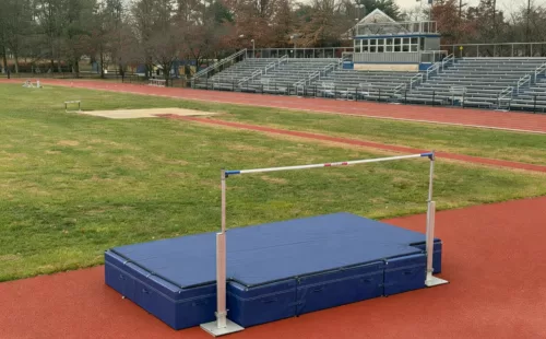 High jump landing mats with standards and bar set up on an outdoor track field beside bleachers and a running track.