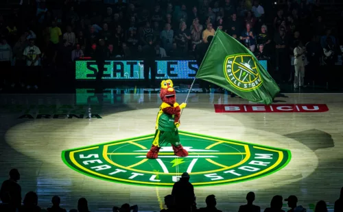 eattle Storm mascot waving an oversized green Seattle Storm spirit flag at center court of Climate Pledge Arena during a WNBA game.