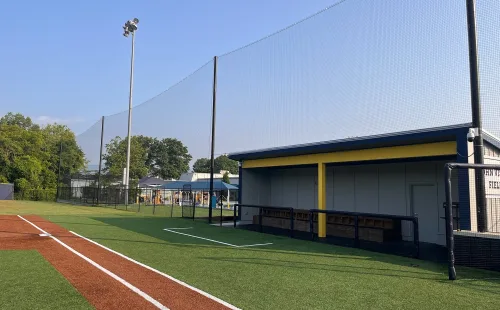 Tall black backstop netting suspended from poles along a turf baseball field's foul line, with a covered dugout to the right.
