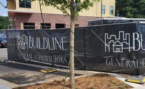 Black mesh construction fence screen printed with the Buildline General Contractors logo on temporary fence panels in front of a building under construction.