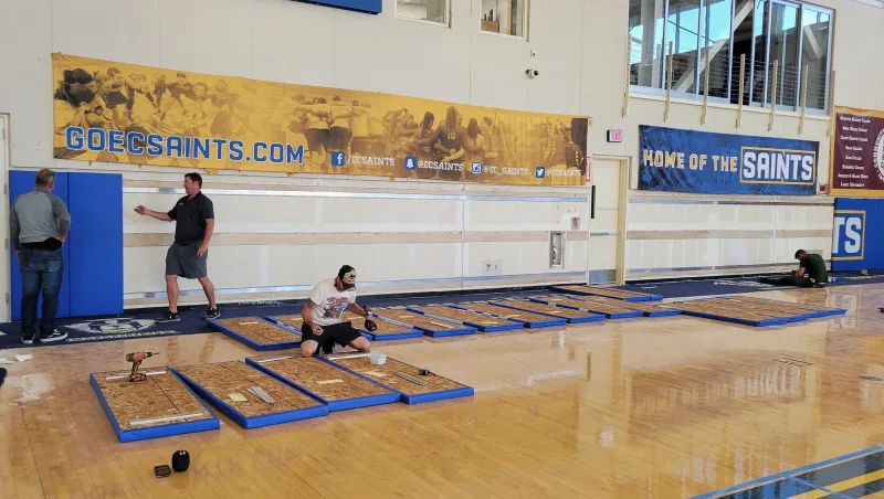 Installers kneeling on a gymnasium floor attaching mounting hardware to the backs of blue woodback gym wall pads, with horizontal furring strips already mounted on the wall behind them